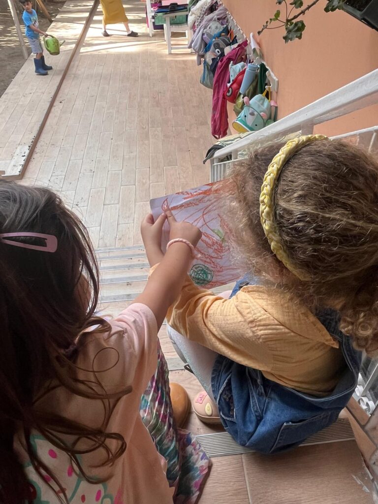 yan yana oturan iki kız çocuğu, birinin elindeki resmi inceler